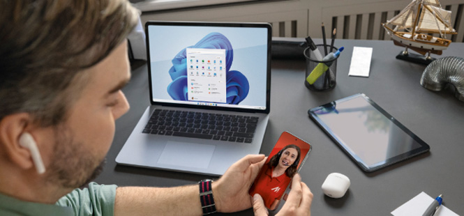 Mężczyzna patrzy na smartfon z wizerunkiem kobiety, obok laptop, tablet i akcesoria biurowe na biurku.