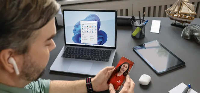 Mężczyzna patrzy na smartfon z wizerunkiem kobiety, obok laptop, tablet i akcesoria biurowe na biurku.