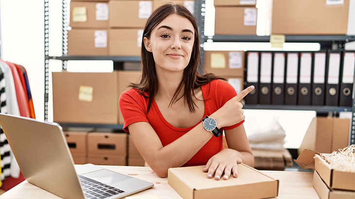 Kobieta przy biurku wskazuje palcem. Za nią regał z kartonami i segregatorami. Obok laptop, pudełka i ubrania.