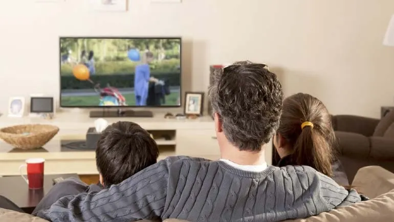Ojciec z dwojgiem dzieci na kanapie przed włączonym 43-calowym TV powieszonym na ścianie salonu. Widok od tyłu.