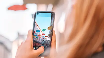 Kobieta trzyma telefon, na ekranie widok ulicy z kolorowymi parasolami. W tle widać inne parasole.