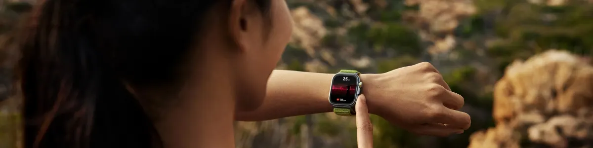 Kobieta patrzy na smartwatch z zielonym paskiem. Na ekranie zegarka widoczny jest wykres i dane. W tle góry i zieleń.