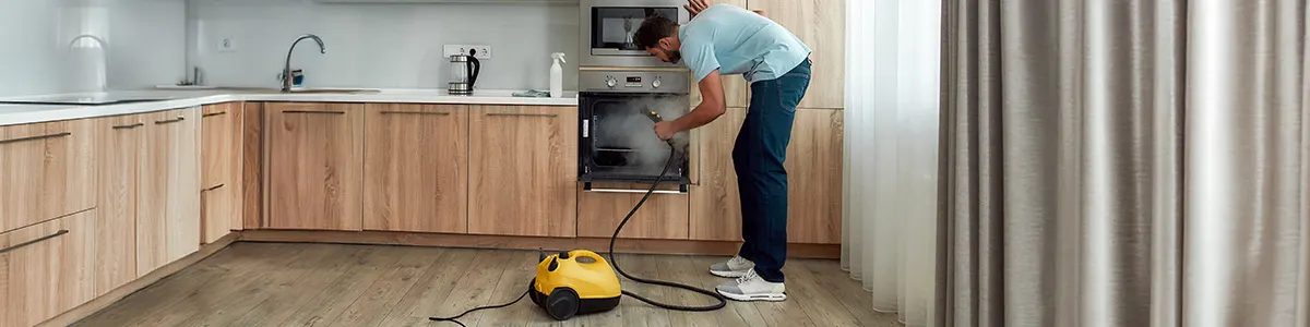 Mężczyzna czyszczący drzwi piekarnika za pomocą żółtej myjki parowej z wężem w nowoczesnej kuchni wykończonej w jasnym drewnie.