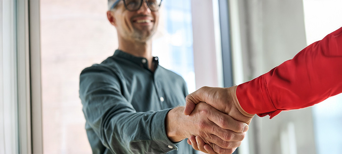 Mężczyzna w szarej koszuli i okularach uśmiecha się, ściskając dłoń osobie w czerwonej koszuli na tle okna.