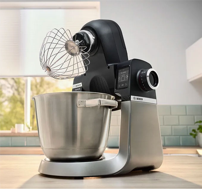 Mikser stojący Bosch z metalową misą i trzepaczką na drewnianym blacie kuchennym, okno w tle.