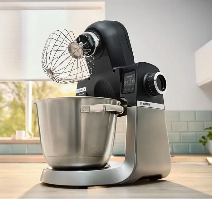 Mikser stojący Bosch z metalową misą i trzepaczką na drewnianym blacie kuchennym, okno w tle.