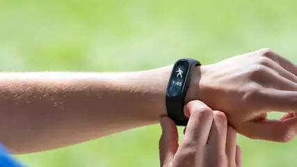 Ręka z czarnym smartbandem na nadgarstku, na ekranie ikona osoby i godzina 1:45, tło zielone.