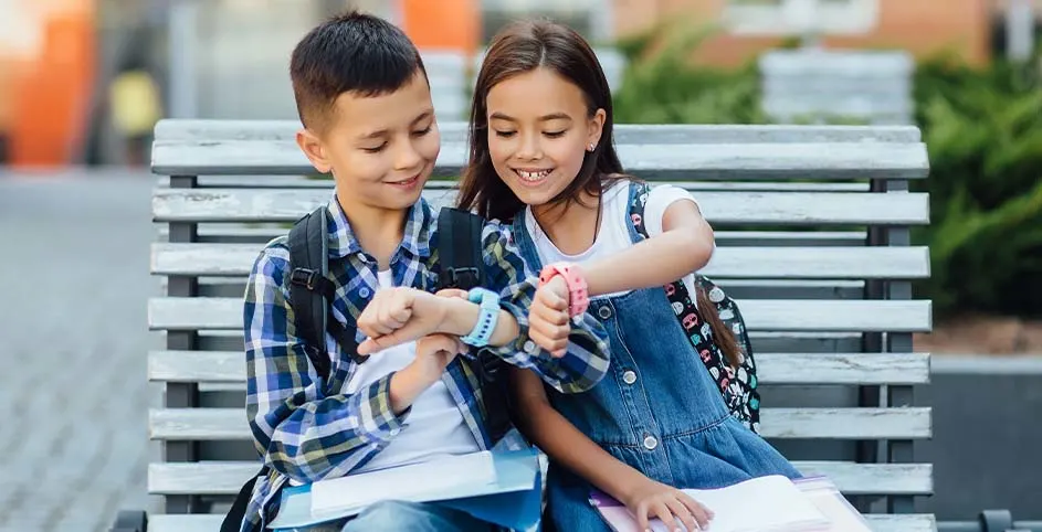 Dzieci siedzą na ławce z plecakami i książkami, patrząc na smartwatche na nadgarstkach. W tle budynek i zieleń.