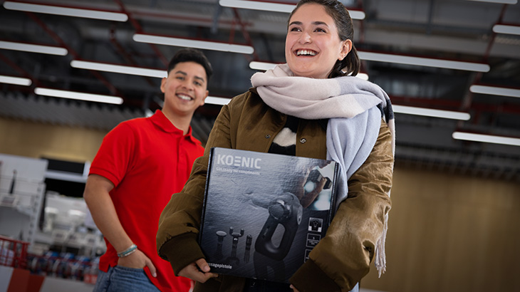 Uśmiechnięta kobieta niosąca zakupiony produkt marki Koenic. W tle uśmiechnięty mężczyzna w czerwonej koszulce