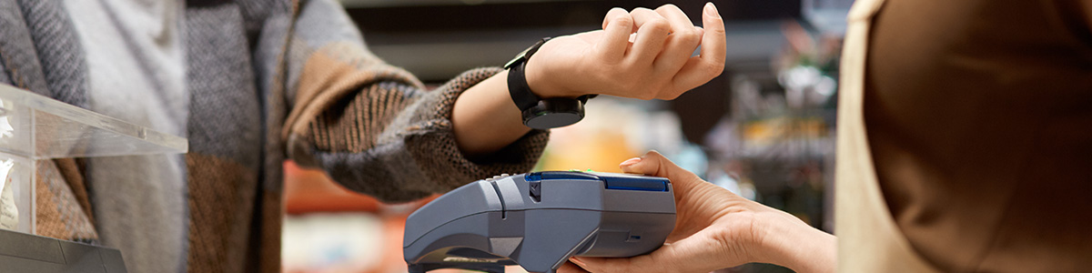 Osoba z zegarkiem zbliża nadgarstek do terminala płatniczego obsługiwanego przez kasjera w sklepie.