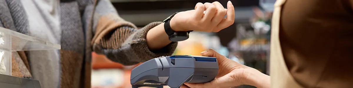 Osoba z zegarkiem zbliża nadgarstek do terminala płatniczego obsługiwanego przez kasjera w sklepie.