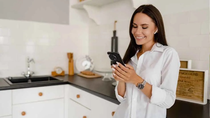 Kobieta w białej koszuli patrzy na telefon w kuchni. W tle białe szafki, czarny blat i zlewozmywak.