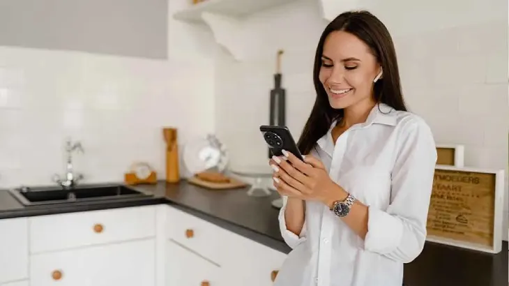 Kobieta w białej koszuli patrzy na telefon w kuchni. W tle białe szafki, czarny blat i zlewozmywak.