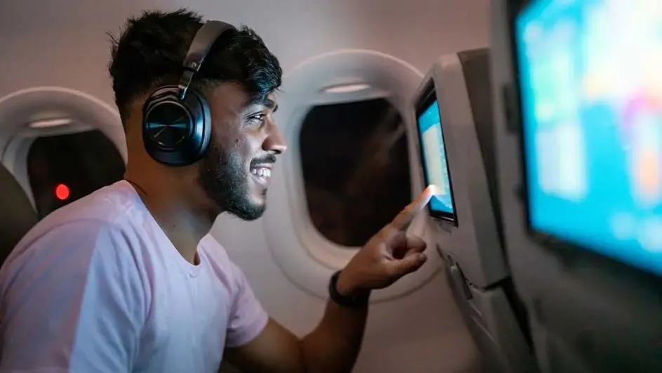 Mężczyzna w słuchawkach w samolocie dotyka ekranu. Za nim okno i zagłówek fotela.