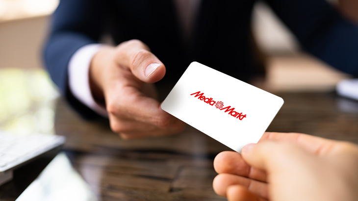 Dwie dłonie wymieniają się białą kartą z czerwonym logo Media Markt na rozmytym tle.