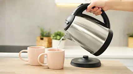 Osoba nalewa wodę z czajnika do różowego kubka na drewnianym blacie. W tle rośliny w doniczkach i wyposażenie kuchni.