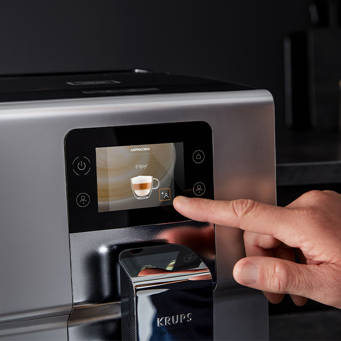 Automat do kawy Krups ze srebrną obudową. Na ekranie dotykowym opcja cappuccino. Palec dotyka ikony na ekranie.