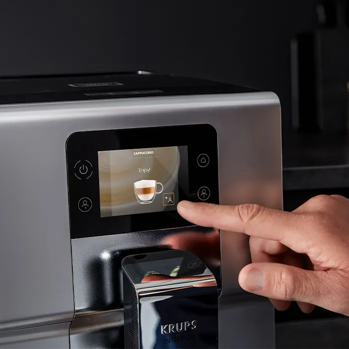 Automat do kawy Krups ze srebrną obudową. Na ekranie dotykowym opcja cappuccino. Palec dotyka ikony na ekranie.