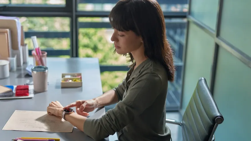 Kobieta siedzi przy biurku, dotykając smartwatcha. Na biurku leżą dokumenty, kubek i przybory do pisania. Za nią widać okno.