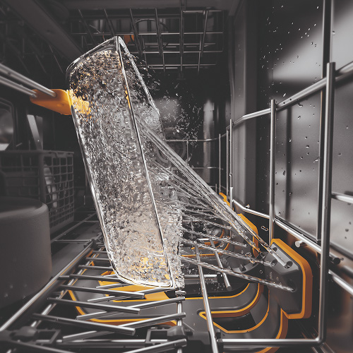 Wnętrze zmywarki z szklanką wypełnioną wodą, umieszczoną na metalowej półce z żółtymi elementami. Woda rozpryskuje się podczas mycia.