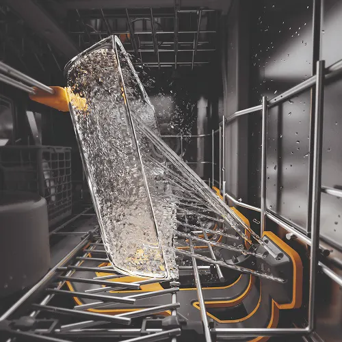 Wnętrze zmywarki z szklanką wypełnioną wodą, umieszczoną na metalowej półce z żółtymi elementami. Woda rozpryskuje się podczas mycia.