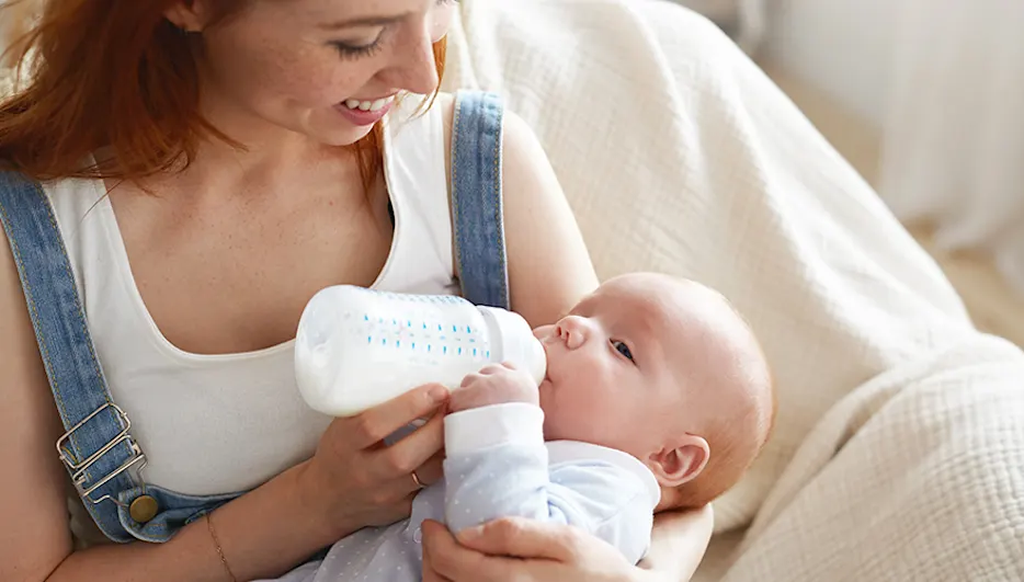 Kobieta karmi butelką niemowlę na jasnym tle. Dziecko trzyma rączkę blisko butelki. Kobieta w białym topie i dżinsowych ogrodniczkach.
