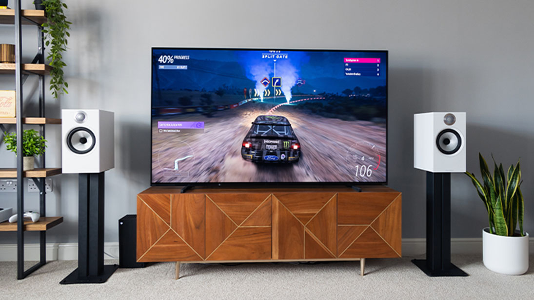 Telewizor 65 cali ustawiony na komodzie, na ekranie gra wyścigowa, po bokach TV białe głośniki.