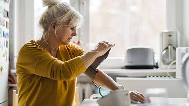 Seniorka w żółtym swetrze mierzy ciśnienie krwi w domu, używając automatycznego ciśnieniomierza na ramię.