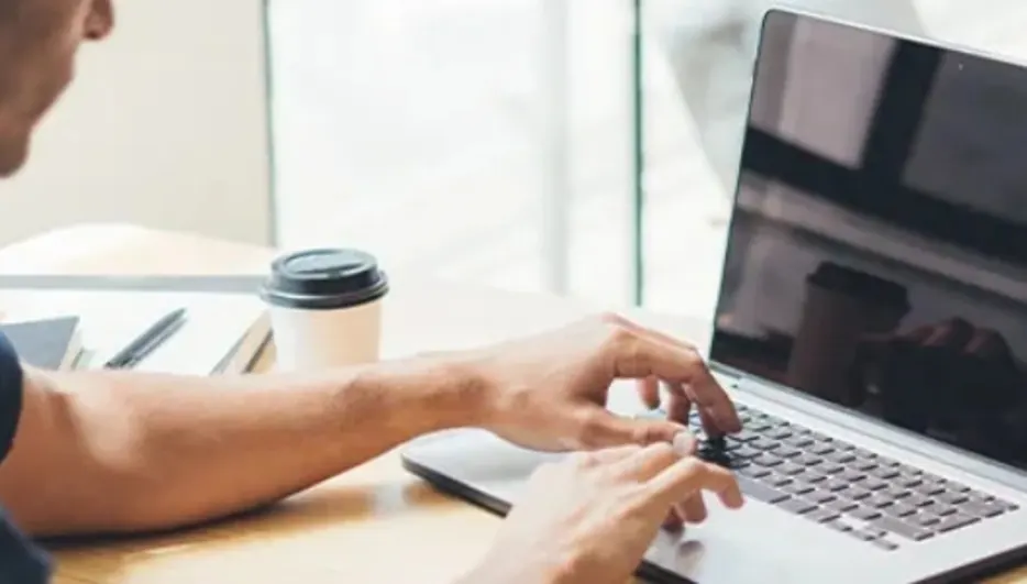 Mężczyzna w niebieskiej koszuli pisze na laptopie przy biurku. Obok stoi kubek z napojem, w tle krzesło.