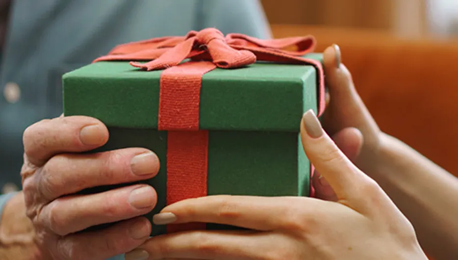 Osoba w niebieskiej koszuli daje zielony prezent z czerwoną wstążką osobie w zielonej koszuli na tle sofy.