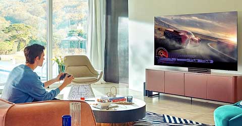 Salon z dużym oknem, mężczyzna gra na konsoli, telewizor pokazuje wyścig samochodowy. Stół, krzesło i szafka pod telewizorem.