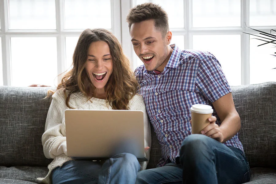 Para siedzi na szarej sofie, patrzy na laptopa z otwartymi ustami. Mężczyzna trzyma kubek. Za nimi okno.