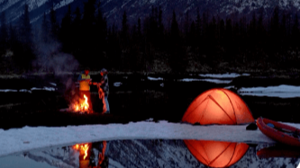 Nocne biwakowanie w górach: dwie osoby stoją przy ognisku obok pomarańczowego namiotu; śnieg, las i skaliste szczyty w tle; odbicie ognia i namiotu w wodzie, po prawej czerwony kajak.