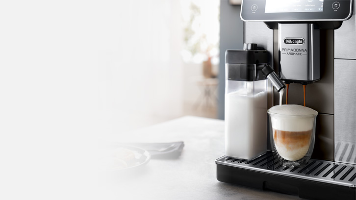 Ekspres do kawy DeLonghi nalewa kawę do szklanki obok pojemnika z mlekiem, na tacy ociekowej.