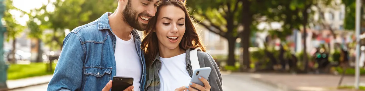 Para trzymająca smartfony stoi na tle parku miejskiego. Mężczyzna w jeansowej koszuli, kobieta w białej koszulce.