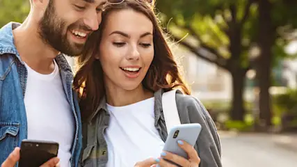 Para trzymająca smartfony stoi na tle parku miejskiego. Mężczyzna w jeansowej koszuli, kobieta w białej koszulce.