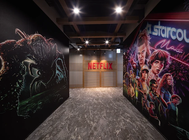 Korytarz z czerwonym logo Netflix w głębi, którego ściany ozdobione są dużymi grafikami przedstawiającymi bohaterów i potwora z serialu "Stranger Things".