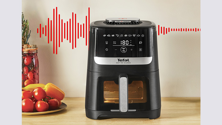 Czarny frytkownica Tefal na blacie, obok słoik z przetworami i talerz z pomidorami i papryką. Czerwone linie na ścianie.