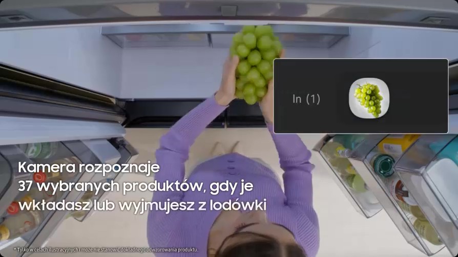 Widok z góry na otwartą lodówkę; osoba wkłada do środka kiść zielonych winogron. Po prawej panel z ikoną talerzyka z winogronami i napisem ‘In (1)’. Na ekranie tekst: ‘Kamera rozpoznaje 37 wybranych produktów, gdy je wkładasz lub wyjmujesz z lodówki’.