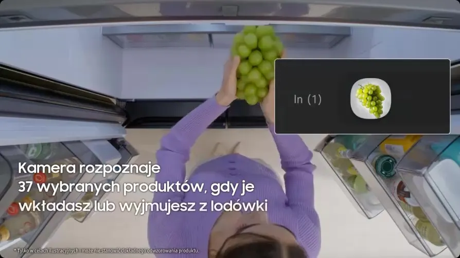 Widok z góry na otwartą lodówkę; osoba wkłada do środka kiść zielonych winogron. Po prawej panel z ikoną talerzyka z winogronami i napisem ‘In (1)’. Na ekranie tekst: ‘Kamera rozpoznaje 37 wybranych produktów, gdy je wkładasz lub wyjmujesz z lodówki’.