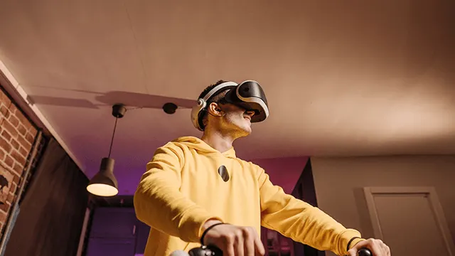 Osoba w żółtej bluzie używa zestawu VR w pokoju z ceglaną ścianą i lampą.