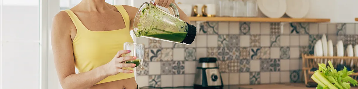 Osoba w żółtej koszulce nalewa zielony koktajl z blendera do szklanki w kuchni. W tle płytki i seler.