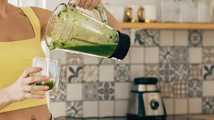 Osoba w żółtej koszulce nalewa zielony koktajl z blendera do szklanki w kuchni. W tle płytki i seler.