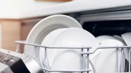 Otwarta zmywarka z metalowym koszem wypełnionym białymi talerzami. Widoczne przyciski na obudowie zmywarki.