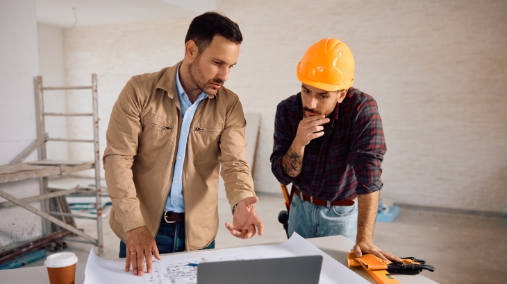 Dwóch mężczyzn przy stole analizuje plany. Jeden w kasku budowlanym, drugi w koszuli i kurtce. W tle drabina i remontowany pokój.