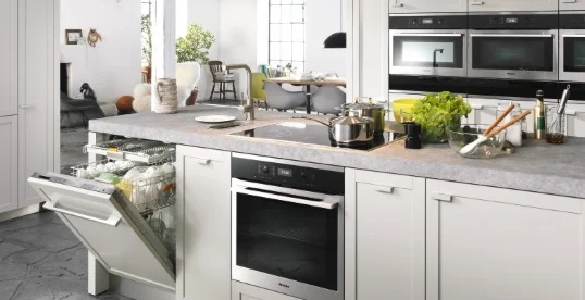 Nowoczesna kuchnia z białymi szafkami, otwartą zmywarką, piekarnikiem i zlewozmywakiem. Na blacie warzywa, naczynia i garnki.