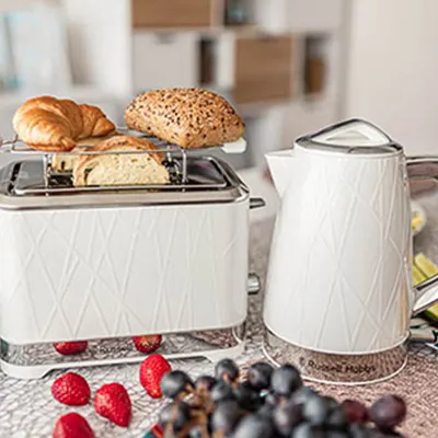 Na stole toster z pieczywem, czajnik, truskawki i winogrona. W tle szafki kuchenne.