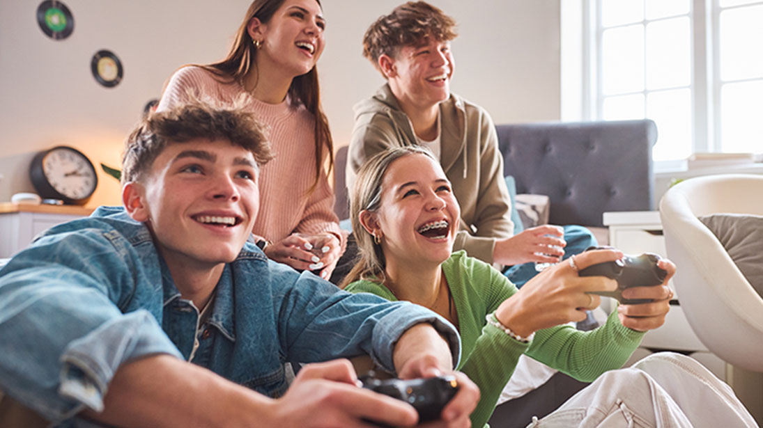 Cztery osoby grają w gry wideo w salonie. Dwie osoby siedzą z przodu i trzymają kontrolery. Dwie osoby stoją za nimi.