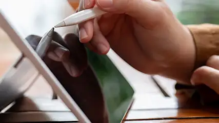 Na drewnianym stole stoi tablet, laptop i kubek. Ręka pisze na ekranie tabletu za pomocą białego rysika.