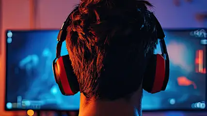Zdjęcie zrobione od pleców osoby, która siedzi przed komputerem, gra w grę i ma śłuchawki na uszach. Na drugim planie komputer i monitor.